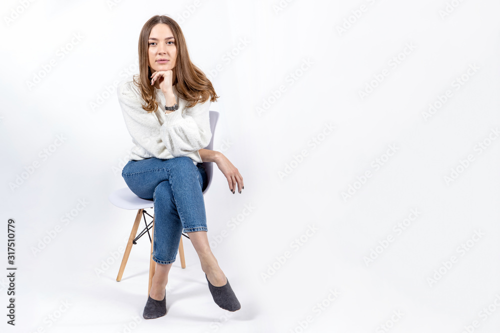 beautiful young girl in jeans and a white sweater sits on a chair propping her head with her hand on a white background, copy space beautiful young girl in jeans and a white sweater sits on a chair propping her head with her hand on a white background, copy space