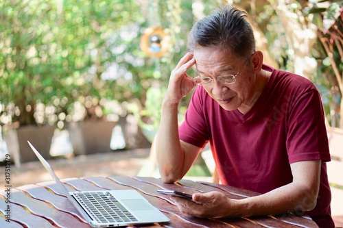 An old asian man in red t shirt and eyeglasses using smart phone and getting headache, with a computer on the table, in the garden