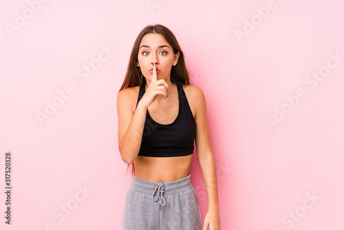 Young caucasian fitness woman posing in a pink background keeping a secret or asking for silence.