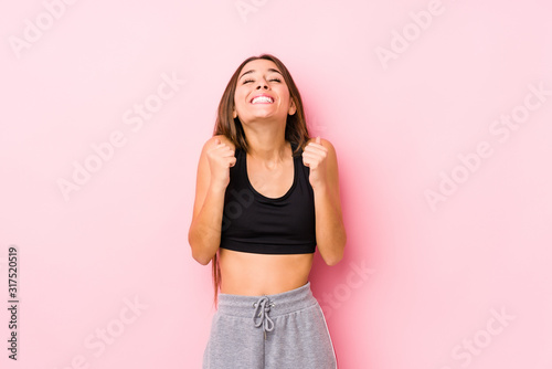 Young caucasian fitness woman posing in a pink background raising fist, feeling happy and successful. Victory concept.