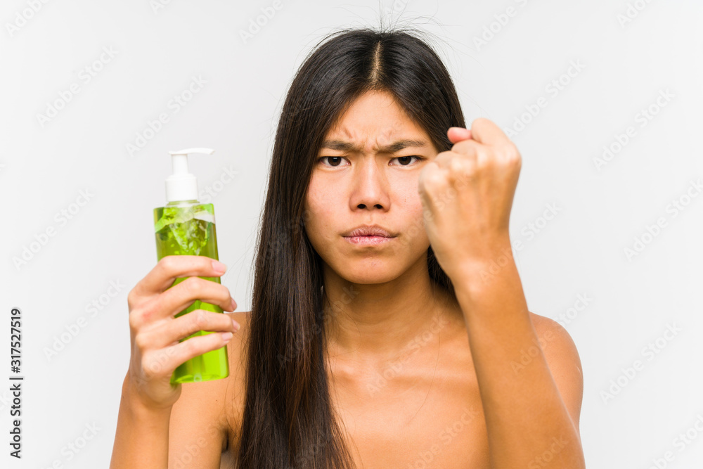 Young chinese woman holding a moisturizer with aloe vera isolated showing fist to camera, aggressive facial expression. Young chinese woman holding a moisturizer with aloe vera isolated showing fist to camera, aggressive facial expression.