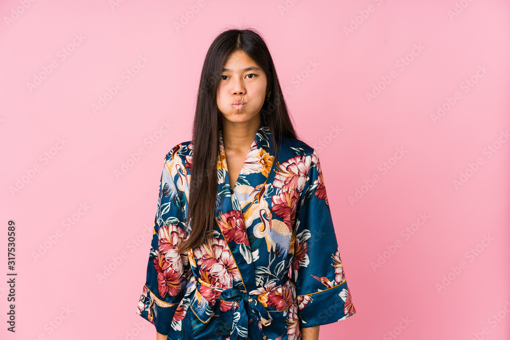 Young asian woman wearing a kimono pajamas blows cheeks, has tired expression. Facial expression concept. Young asian woman wearing a kimono pajamas blows cheeks, has tired expression. Facial expression concept.