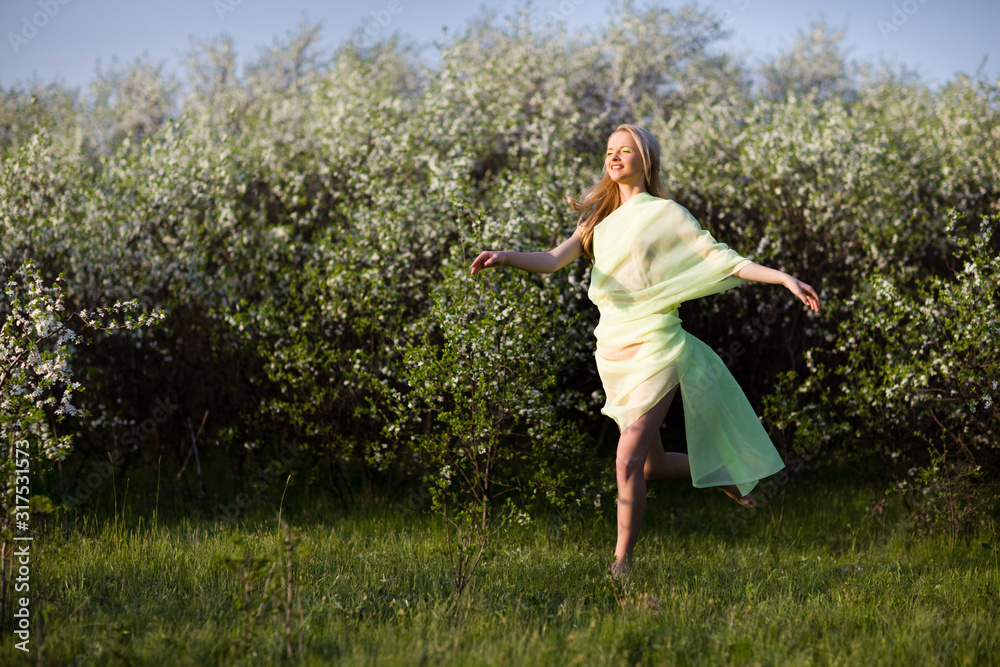 Young beautiful blond smiling woman in yellow transparent cloth jumping on grass over blooming cherry trees background