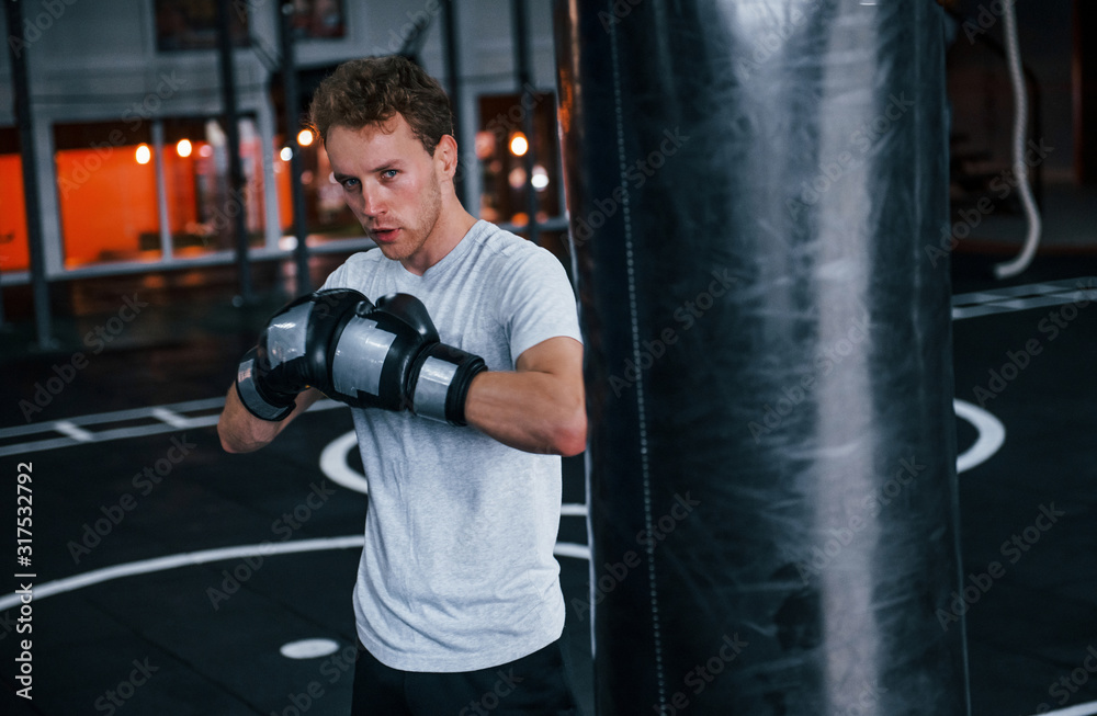 Young man in white shirt and boxing protective gloves doing exercises in gym with pushing bag