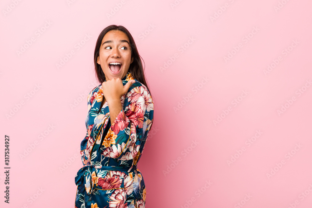 Young indian woman wearing a kimono pajama points with thumb finger away, laughing and carefree. Young indian woman wearing a kimono pajama points with thumb finger away, laughing and carefree.