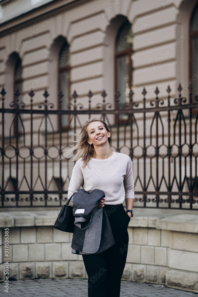 Outdoor photo of blonde lady posing on architecture background in autumn day.Close up fashion street style portrait.wearing dark casual trousers,creamy sweater and gray coat or jacket.Fashion concept.
