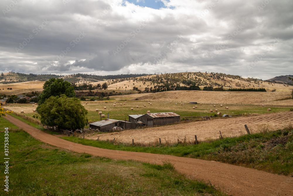 Richmond, Tasmania, Australia - December 13, 2009: Rural landscape with farm under heavy gray cloudscape of forested hills, dirt road, and dry meadows. Corrugated roofs on barns.