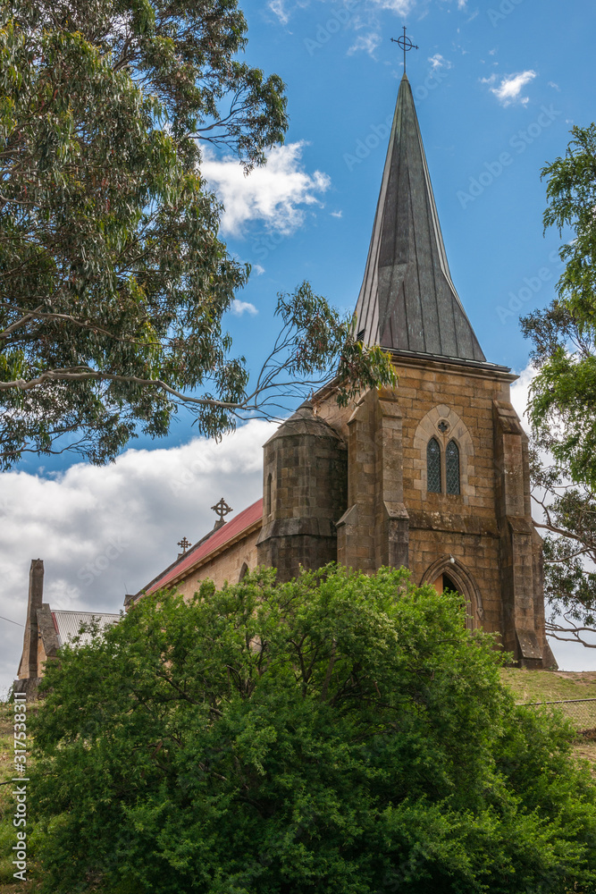 Richmond, Tasmania, Australia - December 13, 2009: Brown stone, red roof and gray spire of Saint Johns Catholic Church. Portrait from behind green bush upon entrance tower under blue sky.
