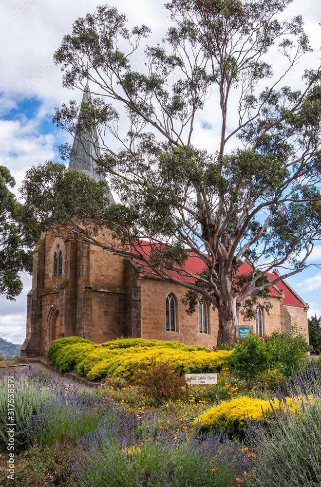 Richmond, Tasmania, Australia - December 13, 2009: Brown stone, red roof and gray spire of Saint Johns Catholic Church. Portrait from side, behind yellow bush and green tree under blue cloudscape.