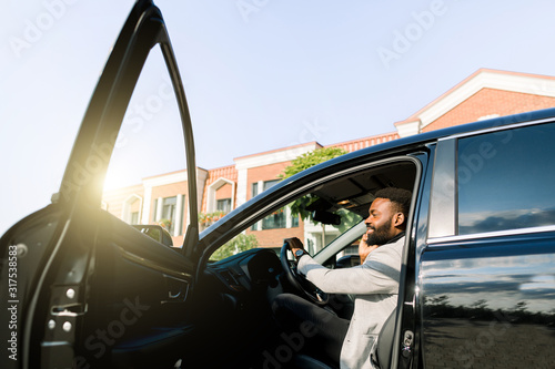 African American male boss businessman using smartphone for making call during while sitting in luxury car with open door, serious man concentrated phoning to partner. Side view © sofiko14