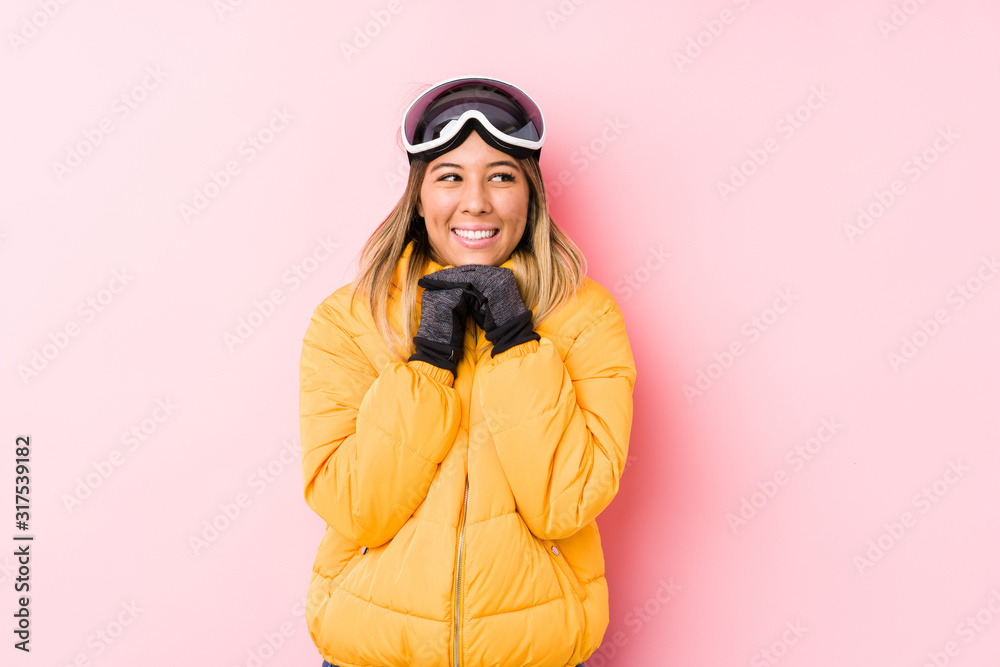 Young caucasian woman wearing a ski clothes in a pink background keeps hands under chin, is looking happily aside.
