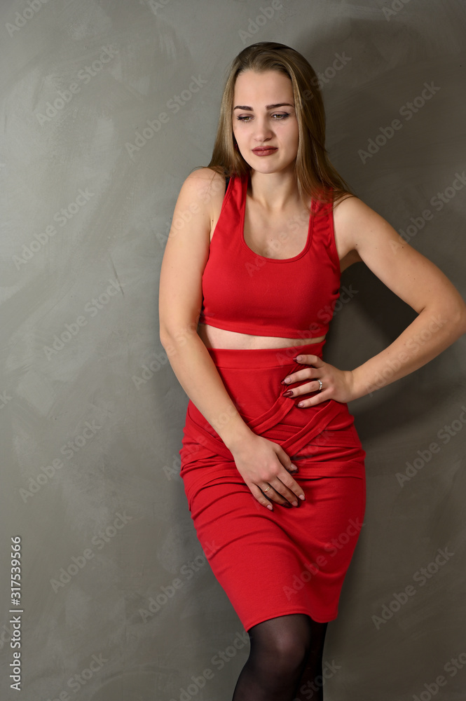 Vertical Photo of a pretty smiling girl with long hair and excellent make-up in a red dress stands next to the wall in various poses. Universal concept female portrait on a gray background. Vertical Photo of a pretty smiling girl with long hair and excellent make-up in a red dress stands next to the wall in various poses. Universal concept female portrait on a gray background.