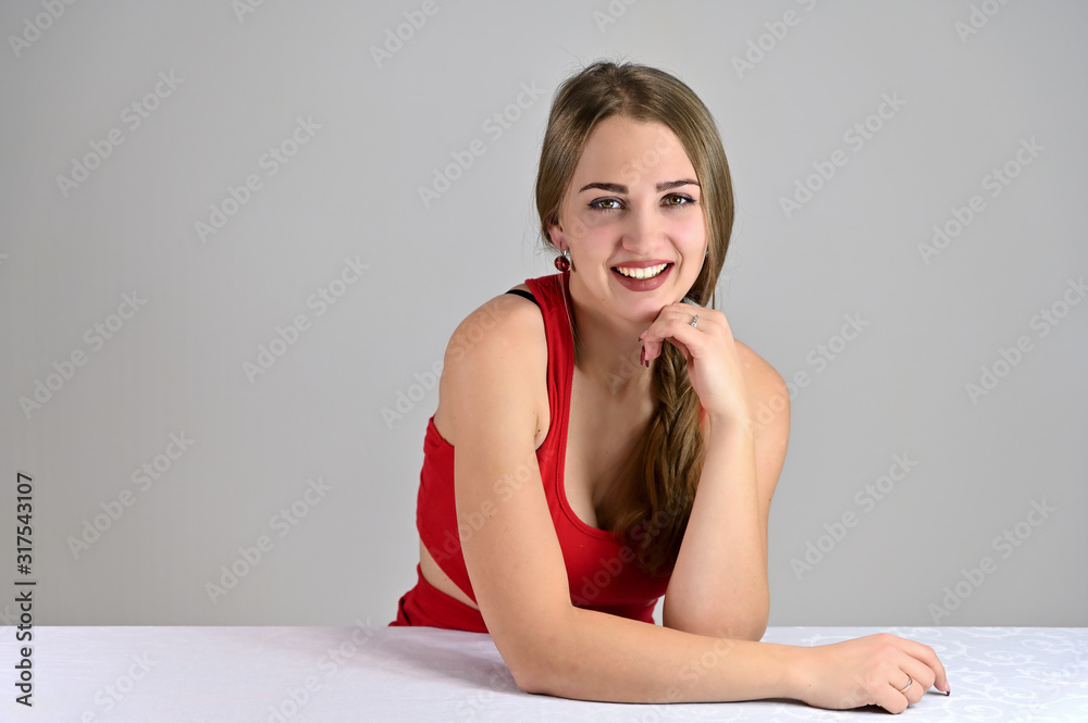 Fototapeta premium Universal concept female portrait on a white background. Horizontal photo of a pretty smiling girl with long hair and great makeup sitting at a white table in the studio.