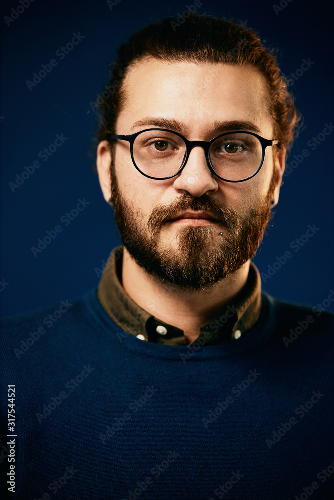 Young attractive caucasian serious bearded hipster with eyeglasses standing in front of blue background. Young attractive caucasian serious bearded hipster with eyeglasses standing in front of blue background.