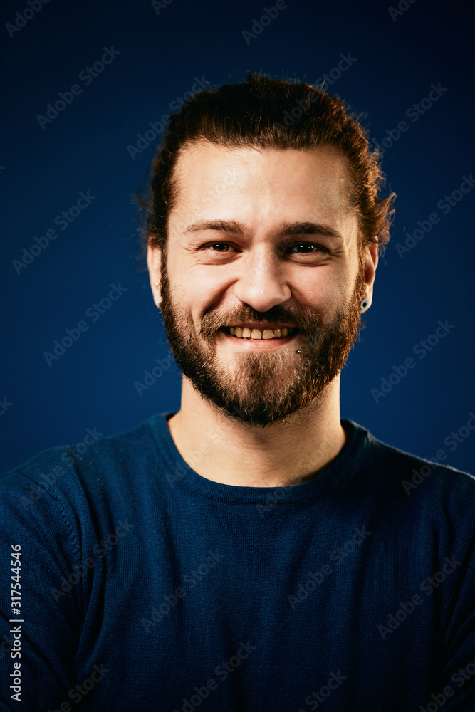 Young handsome smiling bearded caucasian hipster in blue sweater standing in front of blue background and looking at camera.