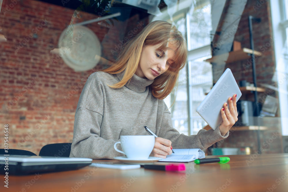 Obraz premium Beautiful caucasian business lady working in office with laptop. Young female model in co-working place makes notes, drinks coffee, performes tasks. Concept of business, finance, freelance, open space