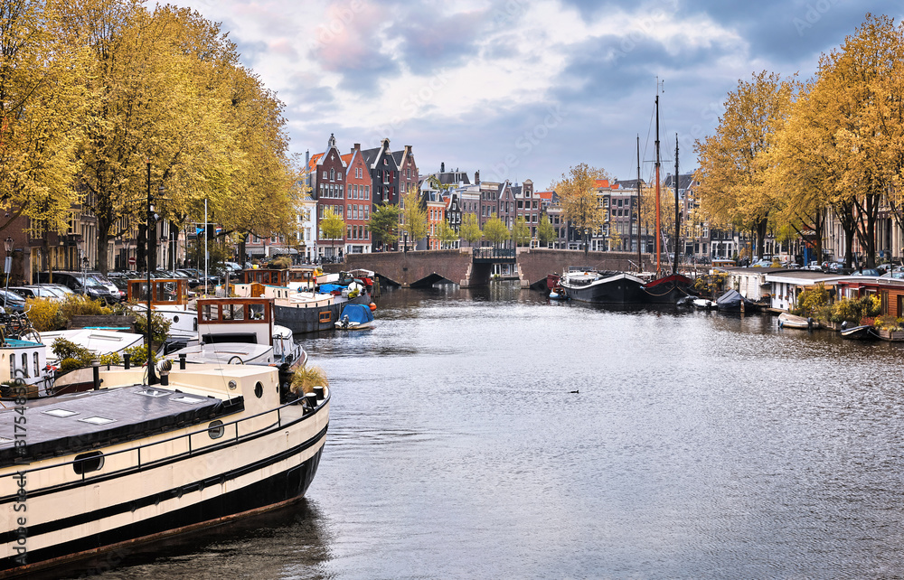 Netherlands Amsterdam. View at river Amstel with boats bridge and traditional houses. Picturesque city landscape. Autumn day in touristic urban with yellow tree.