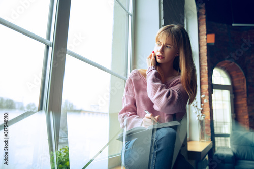 Beautiful caucasian business lady working in office. Young female model in co-working place makes notes, while talking on phone near by window. Concept of business, finance, freelance, open space.
