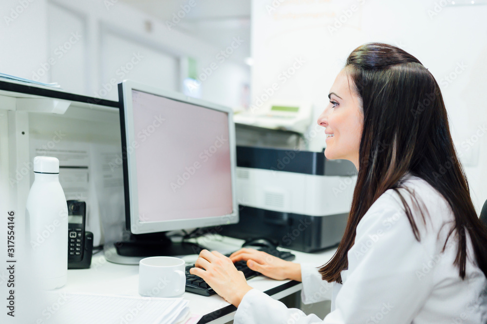 Side view of woman in medical uniform using computer while sitting at table in hospital lab Side view of woman in medical uniform using computer while sitting at table in hospital lab