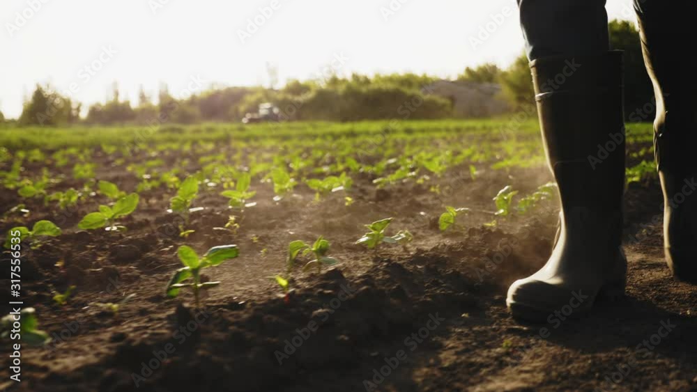 A farmer walks across a field in rubber boots on a blurred background of the tractor in motion.