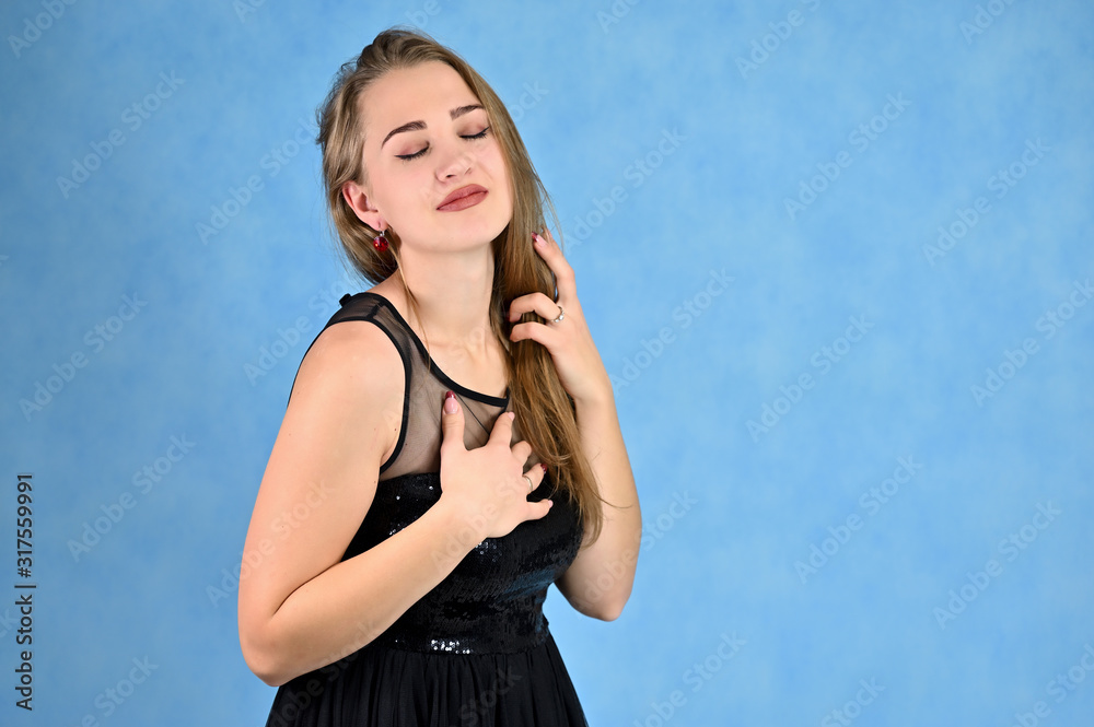 A photo of a pretty smiling girl with long hair and excellent make-up in a black dress stands in different poses. Universal concept horizontal portrait of a woman on a blue background. A photo of a pretty smiling girl with long hair and excellent make-up in a black dress stands in different poses. Universal concept horizontal portrait of a woman on a blue background.