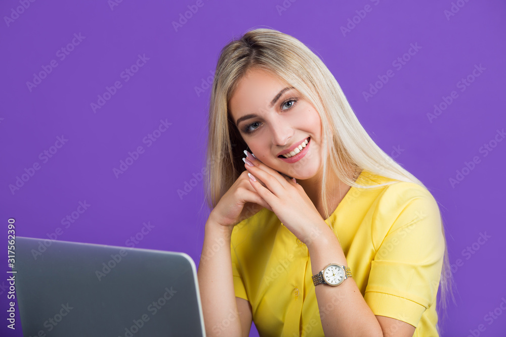 beautiful young woman in a yellow dress on a purple background with her hair sits at a table with a laptop beautiful young woman in a yellow dress on a purple background with her hair sits at a table with a laptop