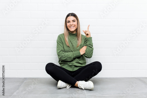 Young caucasian woman sitting on the floor smiling cheerfully pointing with forefinger away.
