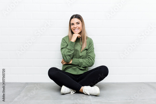 Young caucasian woman sitting on the floor smiling happy and confident, touching chin with hand.