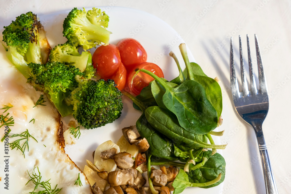 Proper nutrition, healthy eating breakfast of eggs, broccoli, mushrooms, garlic, spinach, dill and small tomatoes close up on white plate with fork lying near. Diet balanced organic food breakfast