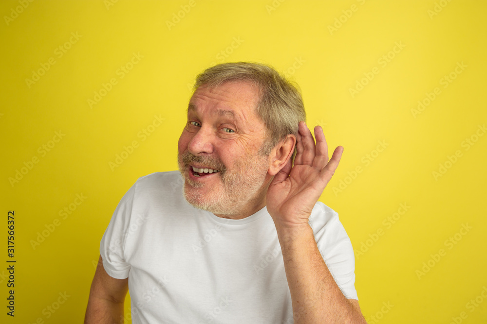 Obraz premium Listening to secret. Caucasian man portrait isolated on yellow studio background. Beautiful male model in white shirt posing. Concept of human emotions, facial expression, sales, ad. Copyspace.
