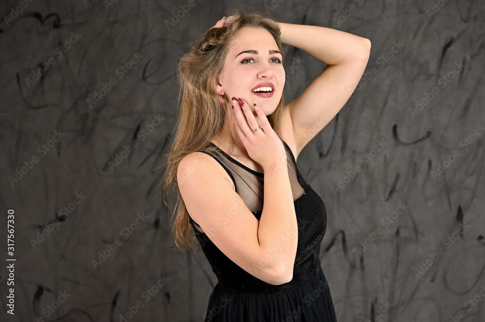 Naklejka premium A photo of a pretty smiling girl with long hair and excellent make-up in a black dress stands in different poses. Universal concept of horizontal female portrait on gray alternative background.
