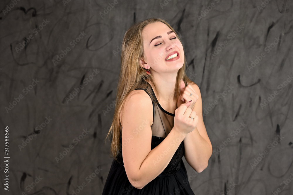Fototapeta premium A photo of a pretty smiling girl with long hair and excellent make-up in a black dress stands in different poses. Universal concept of horizontal female portrait on gray alternative background.