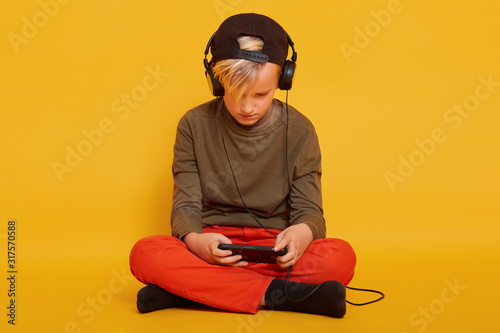 Foto Horizontal studio photo of busy joyful kid having headphones connected to smartphone, playing games, looking at screen attentively, crossing legs, sitting on floor isolated over yellow background