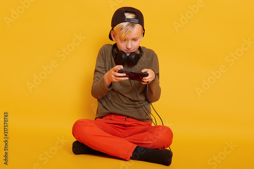 Photo of busy focused blond haired boy looking at smartphone screen attentively, playing games, being enthuthiastic, having headphones around neck, being busy. Children and relaxation concept. © sementsova321