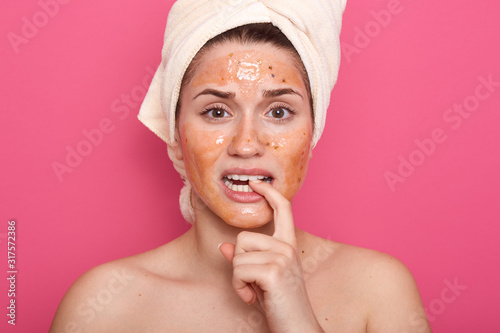 Indoor shot of girl with white towel on her head posing with bare shoulders, female with cosmetic scrab on face. Beauty treatments at home in morning, stands biting her finger against studio wall.