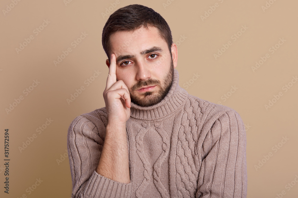 Studio shot of serious unshaven man with beard and mustache, keeps index finger on temple, wearing warm knitted sweater, posing isolated over beige background, trying to remember information.