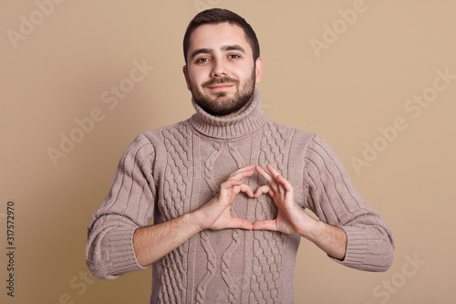 Horizontal shot of handsome male showing heart shape posing isolated over beige background, looking directly at camera, wearing warm knitted sweater, unshaven guy expressing love. People concept.