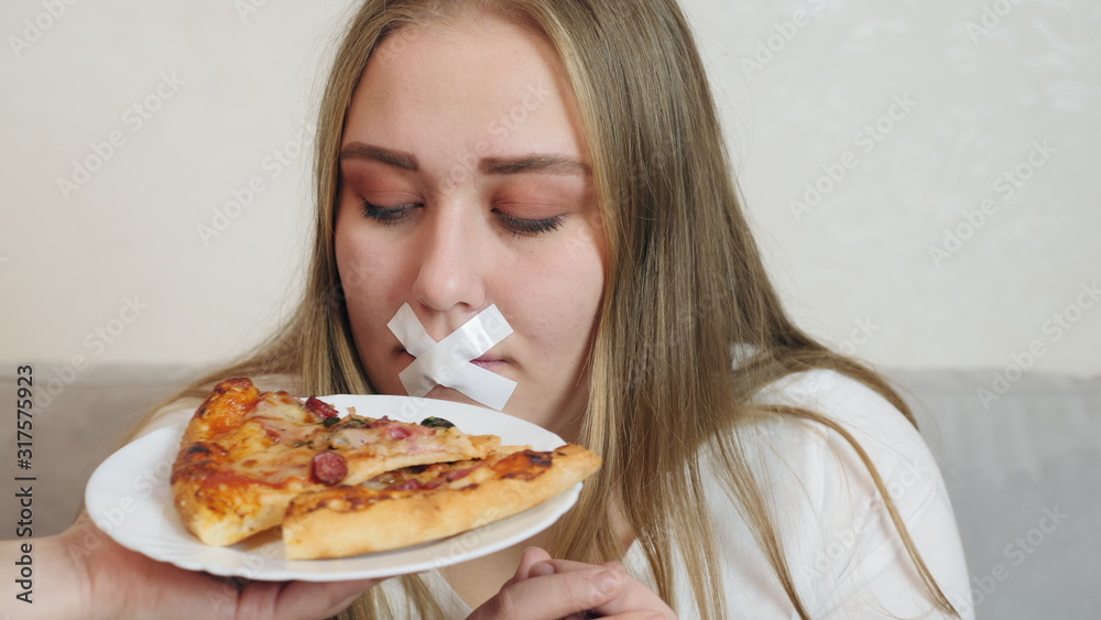 Young woman with white duct tape over her mouth, preventing her to eat junk food. The girl is offered to smell a delicious piece of pizza on a plate. Young woman with white duct tape over her mouth, preventing her to eat junk food. The girl is offered to smell a delicious piece of pizza on a plate.