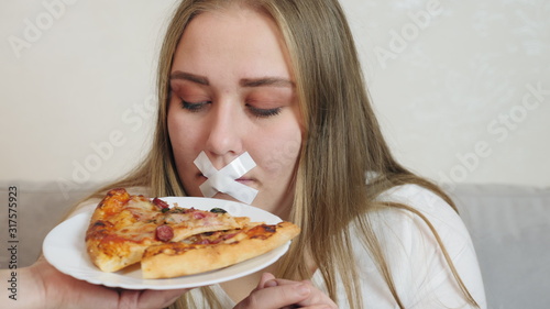 Young woman with white duct tape over her mouth, preventing her to eat junk food. The girl is offered to smell a delicious piece of pizza on a plate.