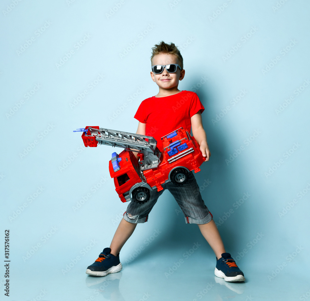 Positive boy in bright stylish casual clothing and sunglasses standing and holding toy fire engine in hands over blue background Positive boy in bright stylish casual clothing and sunglasses standing and holding toy fire engine in hands over blue background