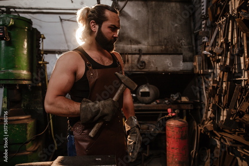 Brutal muscular artisan blacksmith standing in the workshop holding a hammer in his hand in front of a wall of tools