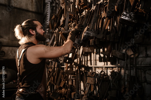 Brutal muscular artisan blacksmith standing in the workshop selects a tool for forging iron