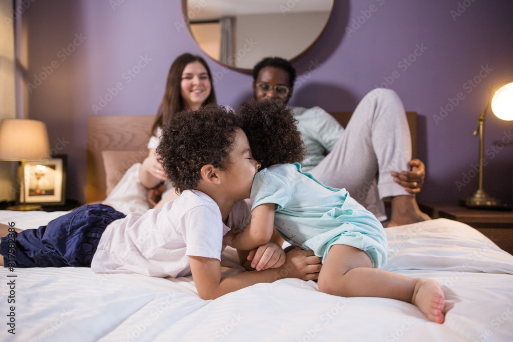 Happy ethnic family with two children sitting on the bed in the bedroom. Little brother and little sister play in the foreground Happy ethnic family with two children sitting on the bed in the bedroom. Little brother and little sister play in the foreground