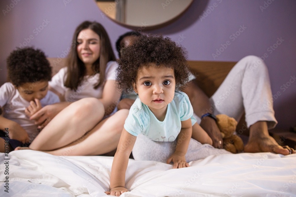 Happy ethnic family lying on the bed in the bedroom. Little sister in the foreground crawling forward