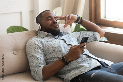 Obraz na plátně African guy holding smartphone closed eyes listen music on headphones