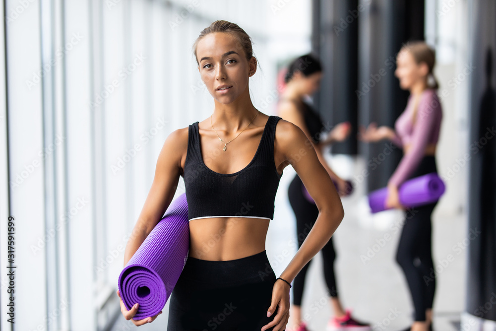 Young fitness woman holding mat in front of two fit girlfriends standing in gym Young fitness woman holding mat in front of two fit girlfriends standing in gym