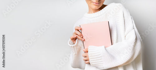 Close view of woman in white woolen sweater holding a book with empty pink cover in hands. Free space for your mock up of reading book concept background.