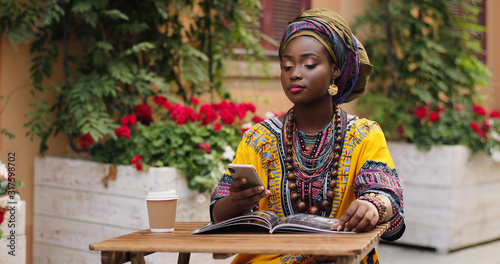 African pretty young woman in the traditional outfit talking on the phone cheerfully while sitting at the table wit magazine and coffee in the beautiful courtyard. © VAKSMANV