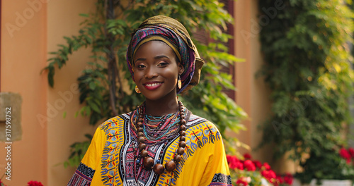 Portrait shot of the beautiful and happy Afrcan woman in the traditional outfit standing in the cozy courtyard with flowers and smiling to the camera. Outdoor. photo