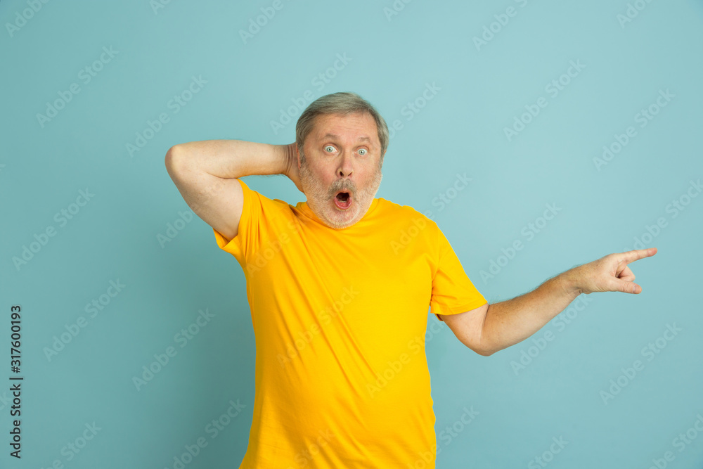 Fototapeta premium Shpcked pointing at side. Caucasian man portrait isolated on blue studio background. Beautiful male model in yellow shirt posing. Concept of human emotions, facial expression, sales, ad. Copyspace.